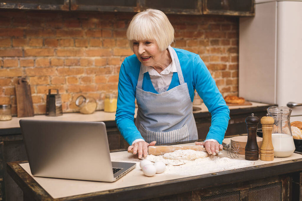 Attractive senior aged woman is cooking on kitchen. Grandmother making tasty baking. Using laptop. - Photo, Image