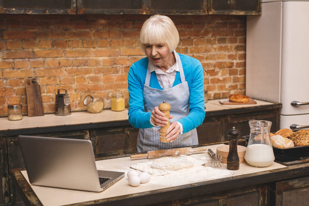 Atractiva anciana mayor está cocinando en la cocina. Abuela haciendo sabrosa cocción. Uso de ordenador portátil
. - Foto, Imagen