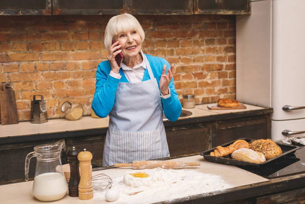Attraente anziana donna sta cucinando in cucina. Nonna che prepara gustosi dolci. Uso del telefono
. - Foto, immagini