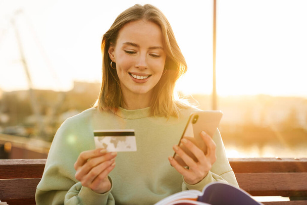 Portrait of smiling young woman holding cellphone and credit card while sitting on bench outdoors - Foto, Bild