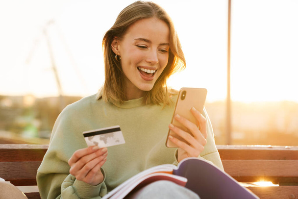 Portrait of excited young woman holding cellphone and credit card while sitting on bench outdoors - Foto, Bild