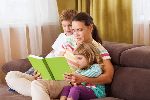 mother reads the book to two of her kids the book on a sofa. The happy family spends time together at home. - Fotó, kép
