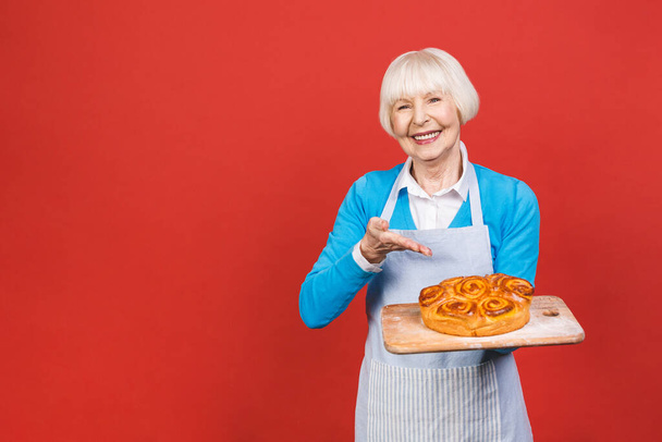 Porträt von ziemlich charmant fröhliche ältere Frau mit Falten zeigt gestikulierenden süßen hausgemachten Kuchen leckeres Brötchen isoliert auf rotem Hintergrund, Spezialität des Hauses. - Foto, Bild