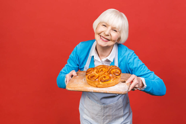 Portrait of pretty charming cheerful senior aged woman with wrinkle showing gesturing sweet homemade pie tasty bun isolated on red background, specialty of the house. - Photo, image