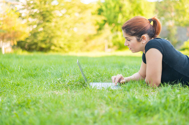 A woman lies on her stomach in a park and types on a laptop. Female freelancer works remotely outdoors. Girl is studying on a computer on the lawn. - Photo, Image