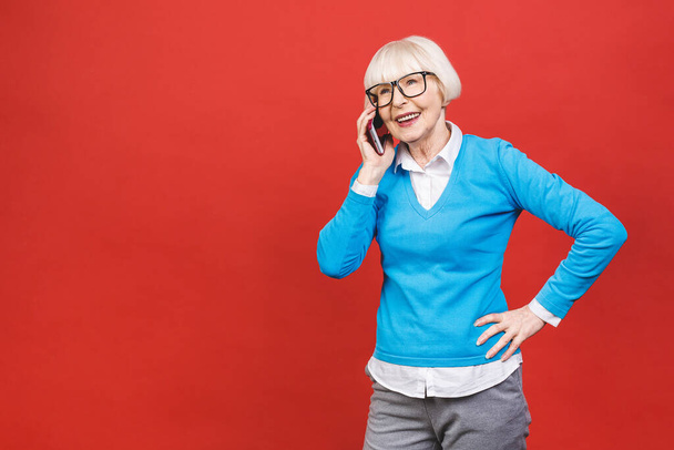 Telefoongesprek. Positieve oudere vrouw glimlachen terwijl het praten op de telefoon geïsoleerd over rode achtergrond. - Foto, afbeelding