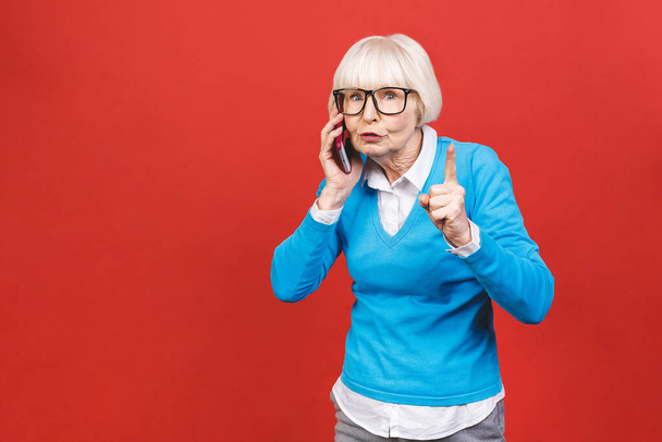 Θυμωμένη ηλικιωμένη γυναίκα μιλάει τηλέφωνο απομονωμένο σε κόκκινο φόντο.  - Φωτογραφία, εικόνα