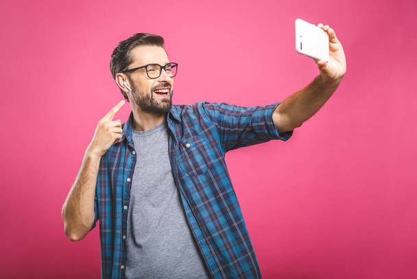 Adoro i selfie! Bel giovanotto in camicia che tiene la macchina fotografica e fa selfie e sorride mentre sta in piedi sullo sfondo rosa. Ascoltare musica con le cuffie
. - Foto, immagini