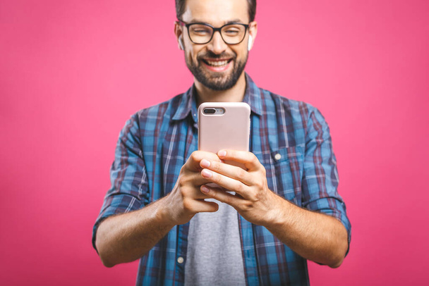 Zawsze w kontakcie. Uśmiechnięty młodzieniec trzymający smartfon i patrzący na niego. Portret szczęśliwego mężczyzny korzystającego z telefonu komórkowego na różowym tle. - Zdjęcie, obraz