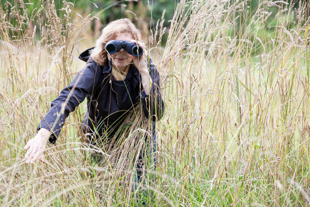 Blond hair European woman spying with binoculars, move to the side high dry grass, black jacket, copyspace - Фото, зображення