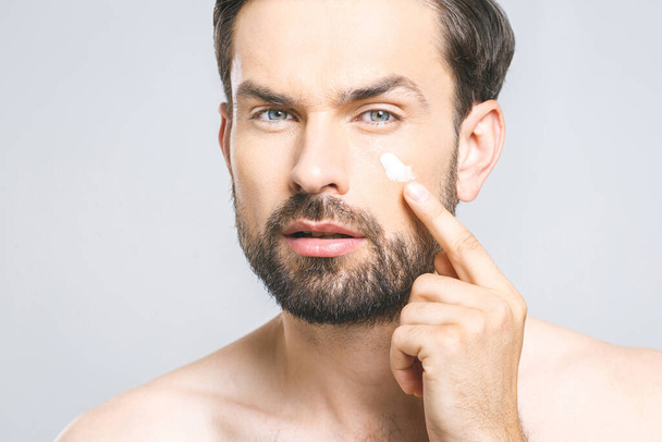 Skin care. Handsome young shirtless man applying cream at his face and looking at himself with smile while standing over gray background and looking at camera. Close-Up. Space for text. - Photo, Image