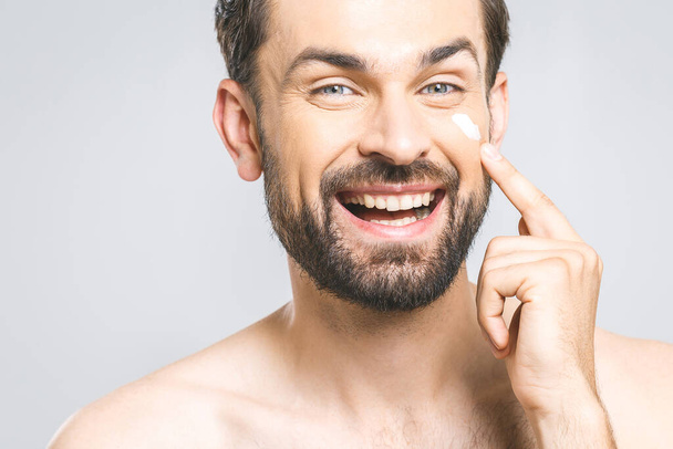 Skin care. Handsome happy young shirtless man applying cream at his face and looking at himself with smile while standing over gray background and looking at camera. Close-Up. Space for text. - Fotó, kép