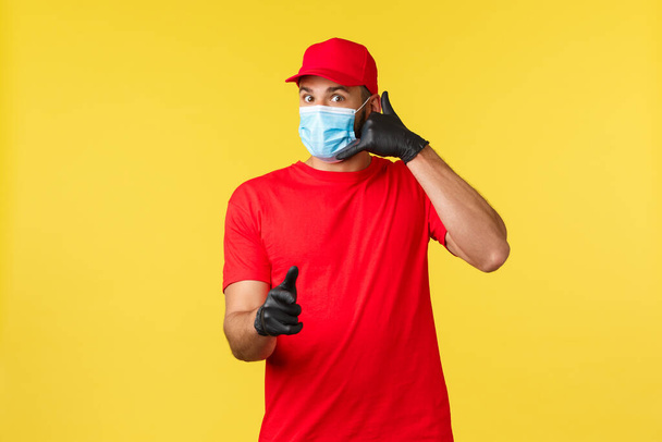 Express delivery during pandemic, covid-19, safe shipping, online shopping concept. Handsome courier in red uniform, medical mask and gloves, pointing client and make phone call sign - Photo, Image