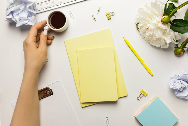 Bürotisch von oben und Frauenhand. Arbeitsplatz mit Klemmbrett, gelben Notizbüchern, Bürobedarf, Pfingstrosenblume und Kaffeetasse, auf weißem Hintergrund. - Foto, Bild