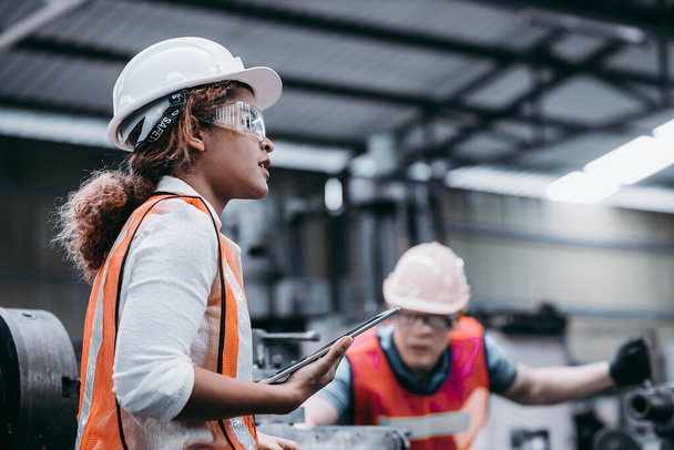 Γυναίκα μηχανικός της βιομηχανίας φορώντας ένα λευκό κράνος, ενώ στέκεται σε ένα βαρύ βιομηχανικό εργοστάσιο πίσω της μιλάμε με τους εργαζόμενους, Διάφορα μεταλλικά μέρη του έργου - Φωτογραφία, εικόνα