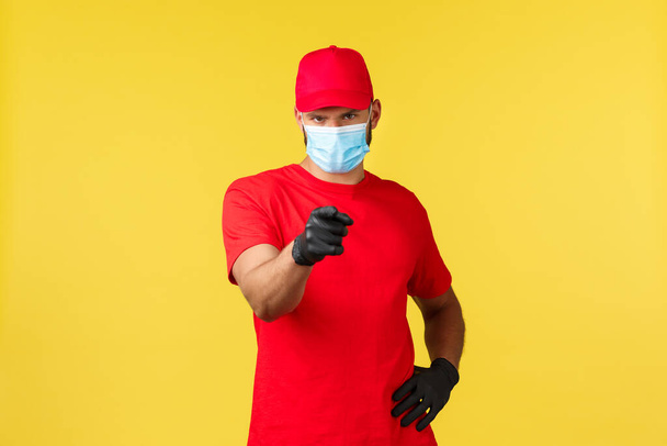 Express delivery during pandemic, covid-19, safe shipping, online shopping concept. Confident serious courier in medical mask and gloves, pointing finger at camera, yellow background - Photo, Image
