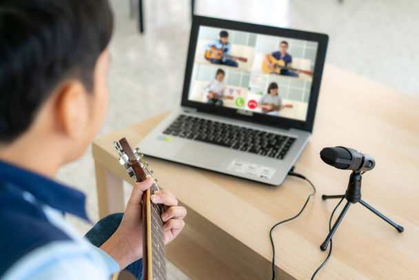 Asiatique garçon jouer guitare acoustique virtuelle happy hour réunion pour jouer de la musique en ligne avec un ami en vidéoconférence avec ordinateur portable pour une réunion en ligne en appel vidéo pour la distance sociale
. - Photo, image