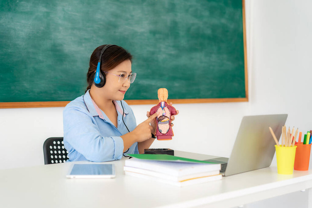 Asyalı kadın öğretmen video konferansında sınıfta karatahta ile e-bilgisayar kullanarak fizyoloji dersi veriyor. Evde eğitim ve uzak öğrenim, çevrimiçi, eğitim ve İnternet. - Fotoğraf, Görsel