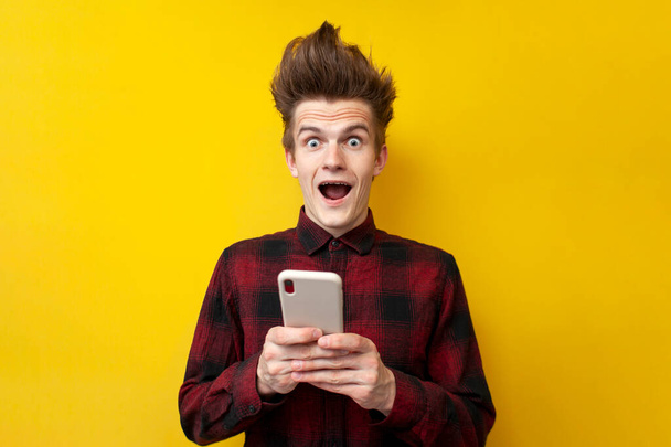 młody zszokowany facet z zabawną fryzurę używać smartfona na żółtym odizolowanym tle, hipster trzyma telefon i jest zaskoczony - Zdjęcie, obraz