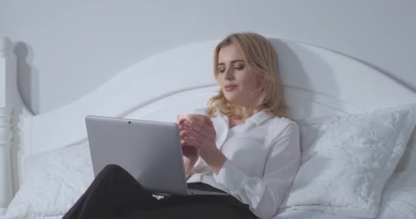 Elegant businesswoman working on laptop enjoys a cup of hot tea before bedtime after a long hard day. Business woman relaxes in bed after a long flight. Business concept. Prores 422 - Záběry, video