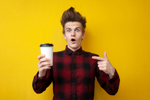 mladý šokovaný chlap pije osvěžující kávu a je překvapen žlutým izolovaným pozadím, muž pije energetický nápoj - Fotografie, Obrázek