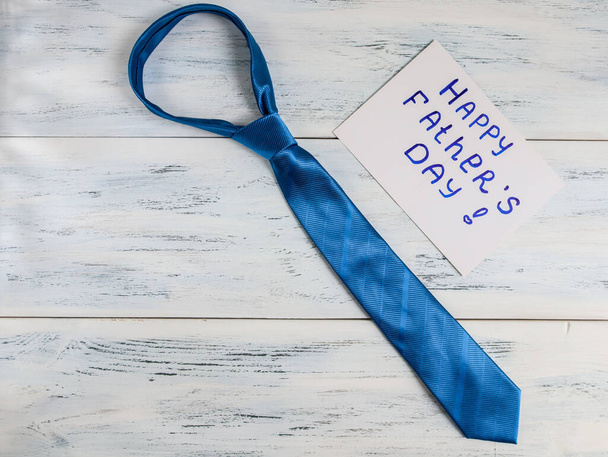 niebieski krawat na drewnianym stole... Niebieski krawat, kartka z napisem "Happy Fathers Day" leżą po prawej stronie na białym drewnianym stole, widok z bliska, z miejscem na tekst po lewej stronie.. - Zdjęcie, obraz