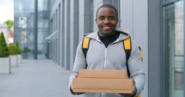 Afroameričtí mladí veselí doručovatelé v batohu dávají balíky kamerám na ulici. Muž kurýr s taškou přinášející krabice pro zákazníka a podání venku. - Záběry, video