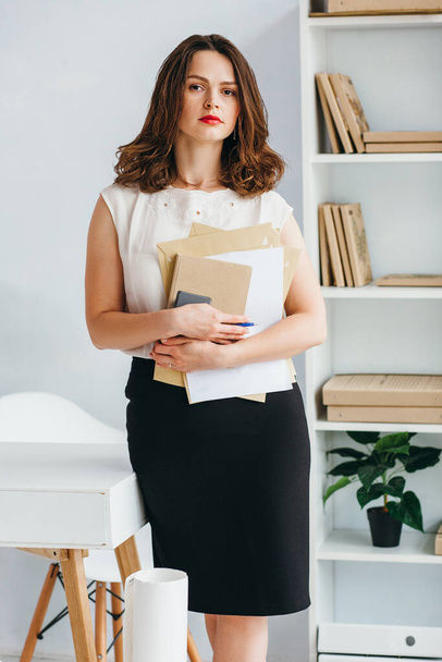 Ένα κορίτσι με άσπρη μπλούζα στέκεται με το τηλέφωνο και τα βιβλία στα χέρια της στο γραφείο. Έχει καστανά μαλλιά κομμένα κάτω από το τετράγωνο..  - Φωτογραφία, εικόνα