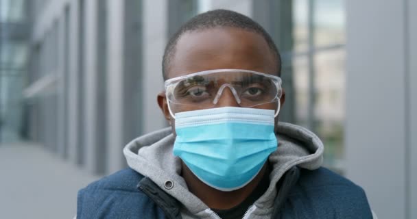 Close-up de cara de jovem homem afro-americano bonito em máscara médica e óculos olhando diretamente para câmera ao ar livre. Retrato de mensageiro masculino na rua durante o coronavírus. Conceito de quarentena
. - Filmagem, Vídeo