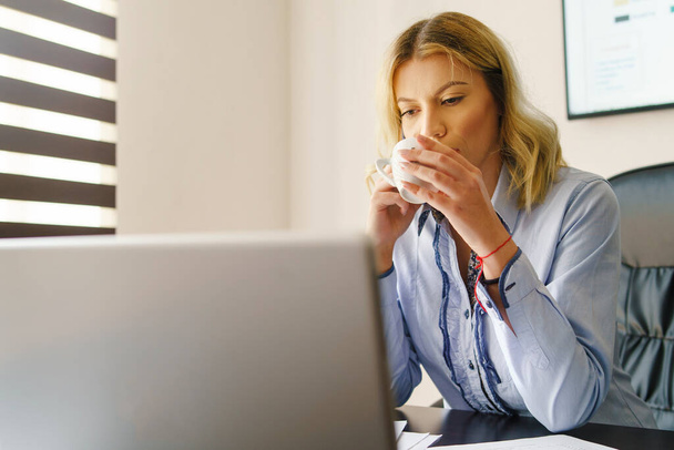 Portret van de jonge volwassen blanke vrouw die op kantoor werkt - Mooi meisje op het werk die een kopje koffie drinkt terwijl ze rapporten leest op laptop - vrouw op het werk die pauze heeft om na te denken over het project - Foto, afbeelding