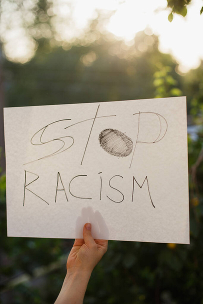 Женщина держит белую бумагу с текстом "Остановите расизм". На фоне деревьев и солнечного света
 - Фото, изображение