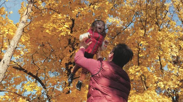 Ο μπαμπάς ξερνάει μια ευτυχισμένη κόρη στον ουρανό το φθινόπωρο στο πάρκο. Ευτυχισμένα οικογενειακά ταξίδια. Πατέρας και μικρό παιδί παίζουν, γελάνε και αγκαλιάζονται. υγιές παιδί στην αγκαλιά του γονέα. Ο μπαμπάς έφυγε. Οικογενειακή έννοια - Φωτογραφία, εικόνα