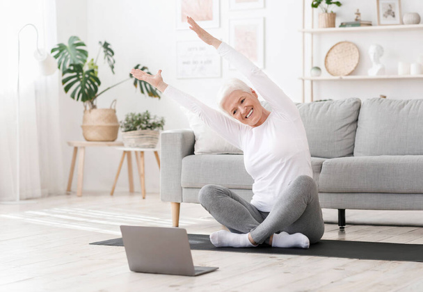 Positive Seniorin dehnt sich vor dem Heimtraining vor Laptop aus - Foto, Bild