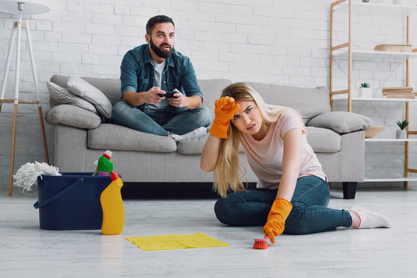 掃除と性差別。男はビデオゲームで楽しみを持っています,彼女の頭に疲れ妻のクリングとハウスキーピングを行う - 写真・画像