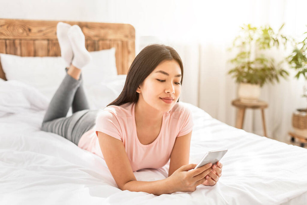 自宅でベッドの上に横たわっているスマートフォンのテキストを持つ中国人女性 - 写真・画像