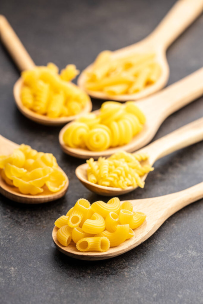 Various types of raw italian pasta in wooden spoons on black table. - Фото, зображення