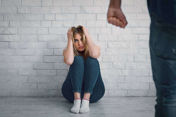 Stop huiselijk geweld. vrouw zit op de vloer, bedekt hoofd met haar handen en kijkt naar man met gebalde vuist - Foto, afbeelding