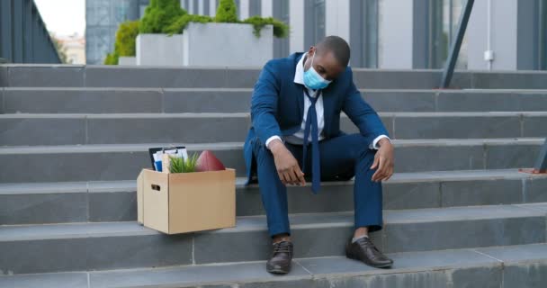 Afro-Amerikaanse trieste zakenman met medisch masker zittend op een trap buiten met doos vol spullen als verloren zaken. Mannelijke kantoormedewerker in wanhoop verloor zijn baan. De werkloosheid neemt toe als gevolg van een pandemie. Ontslagen man - Video