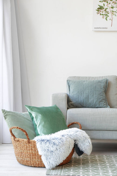 Ψάθινο καλάθι με μαξιλάρια και καρό κοντά στον καναπέ στο δωμάτιο - Φωτογραφία, εικόνα