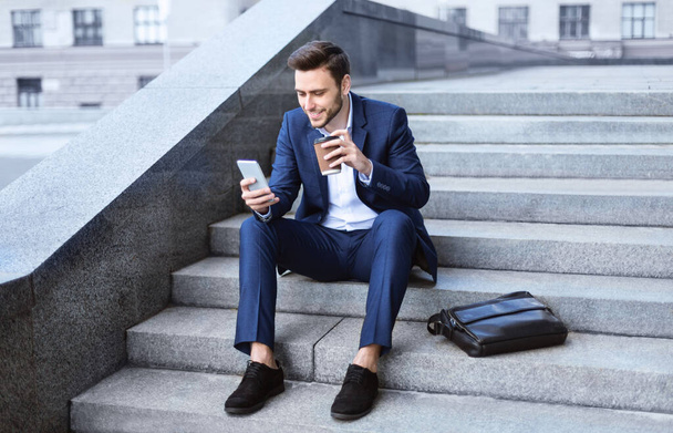 Ευτυχής επιχειρηματίας σε επίσημη φορούν πόσιμο καφέ takeout και περιήγηση δίχτυ μέσω smartphone στις σκάλες κατά τη διάρκεια του γεύματος - Φωτογραφία, εικόνα