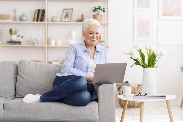 Glimlachende oudere dame ontspannen met laptop op de bank thuis - Foto, afbeelding