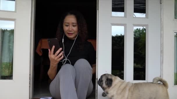 Woman chatting on her cellphone with earphones. Home setting with pug dog. Work at home concept. - Кадри, відео