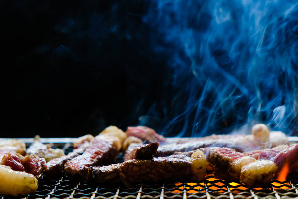 ωμό κρέας μπριζόλα βοείου κρέατος φέτες bbq ψησίματος σε σχάρα ξυλάνθρακα σόμπα με καπνό και φωτιά φλόγα σε μαύρο φόντο. - Φωτογραφία, εικόνα