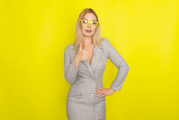 Attractive blonde woman in plaid jacket dress over yellow background wearing yellow glasses - Foto, Imagen