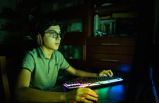 Játék akció közben videojáték ülésen. A fehér fiú hullámos fekete hajjal és szemüveggel az arcát megvilágítja a változó színek a képernyőn a játék során. Játékfülhallgató viselése. - Fotó, kép