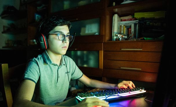 Spielhandlung während einer Videospielsession. Der kaukasische Junge mit den welligen schwarzen Haaren und der Brille lässt sich während des Spiels von den wechselnden Farben des Bildschirms beleuchten. Tragen Sie Gaming-Kopfhörer. - Foto, Bild