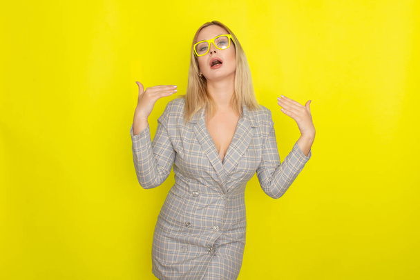 Attractive blonde woman in plaid jacket dress over yellow background wearing yellow glasses - 写真・画像