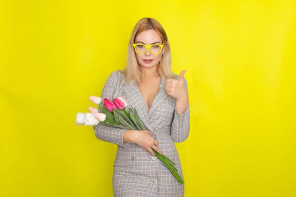 Blonde woman in plaid dress holding tulips bouquet over yellow background - Foto, afbeelding