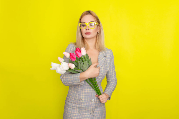 Blonde woman in plaid dress holding tulips bouquet over yellow background - Foto, Bild