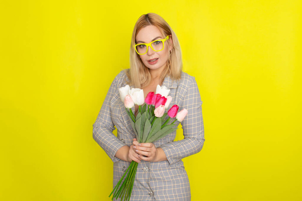 Blonde woman in plaid dress holding tulips bouquet over yellow background - 写真・画像
